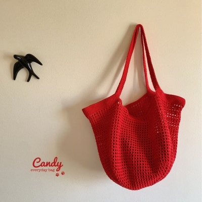 Saco de malha vermelho pendurado em parede creme com gancho negro e texto 'Candy everyday bag'