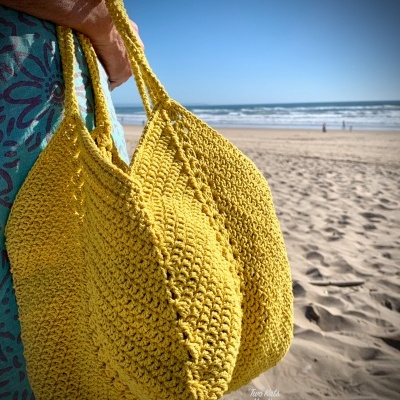 Bolsa de croché amarela na praia, segurada por uma pessoa, com mar ao fundo.