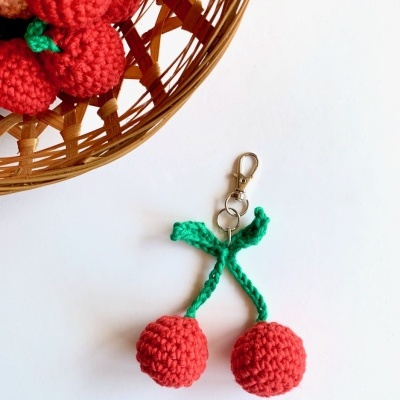 Chaveiro de cerejas de crochê com argola e mosquetão ao lado de cesta de palha com peças iguais