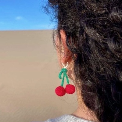 Brincos de cereja tricotados vermelhos e verdes numa orelha com cabelo encaracolado