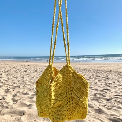 Sacolas amarelas tricotadas seguradas na praia com mar e céu azul ao fundo