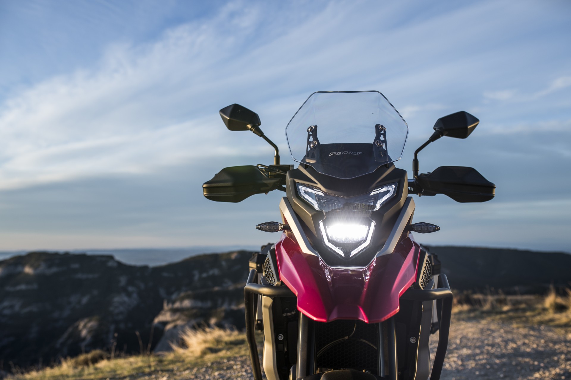 Motociclo vermelho e preto visto de frente com farol aceso em ambiente exterior