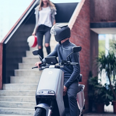 Scooter elétrica cinzenta conduzida por pessoa com capacete preto em área urbana com escadas e parede de tijolos