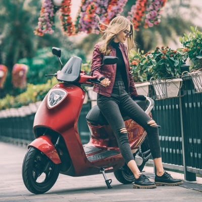 Mulher com jaqueta bordo sentada em scooter vermelho numa calçada com plantas e decoração floral ao fundo