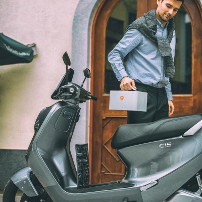 Motociclo elétrico cinzento com homem a segurar caixa branca em frente a porta de madeira