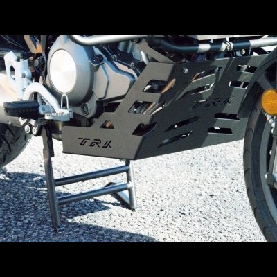 Protector de motor preto com texto TRK numa moto sobre chão de gravilha