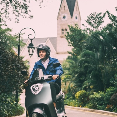 Homem numa scooter elétrica cinza escuro com capacete e casaco azul a conduzir numa estrada arborizada