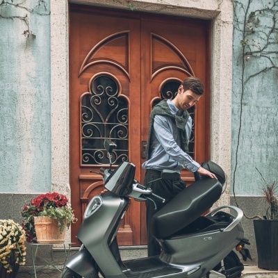 Scooter cinzenta com homem junto a porta de madeira e vasos com plantas
