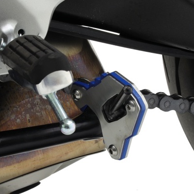 Peça metálica azul e prata ligada à corrente de motocicleta com apoio de borracha preta e parte do escape metálico escurecido