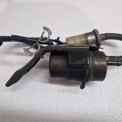 Peça automóvel metálica com tubo preto flexível e suporte metálico sobre superfície branca