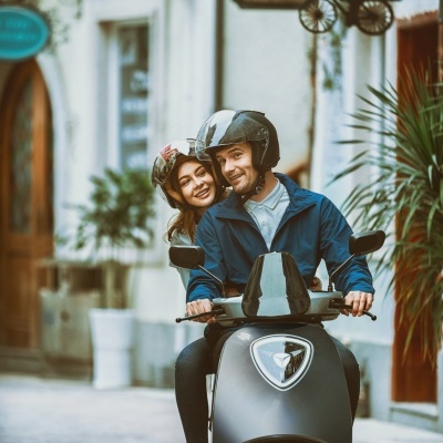 Casal numa scooter preta numa rua com edifícios brancos e plantas