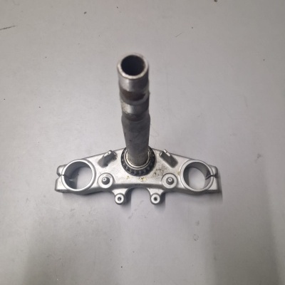 Peça metálica prateada com eixo central e duas aberturas circulares sobre superfície cinzenta