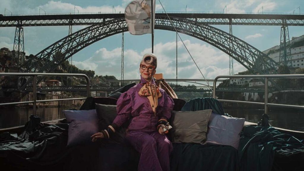 Homem em fato roxo sentado num sofá com almofadas junto à ponte metálica