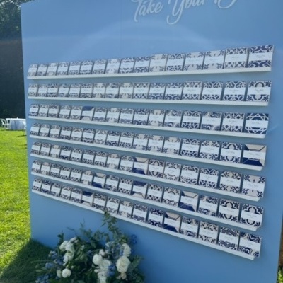 Painel azul claro com cartas azuis e brancas e flores brancas e azuis no chão, ao ar livre