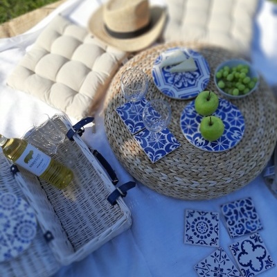 Cesta de piquenique com porcelanas azuis e brancas sobre manta branca com almofadas e chapéu de palha.