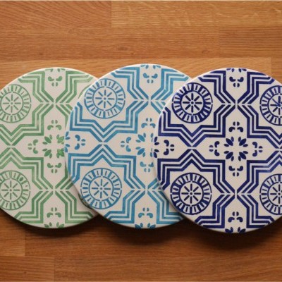 Três bases de copos redondas com padrões geométricos verdes, azuis claras e azuis escuras sobre madeira