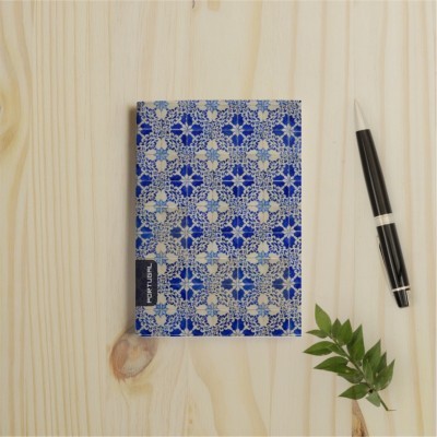 Caderno azul com padrão floral e etiqueta Portugal sobre mesa de madeira, caneta e planta ao lado