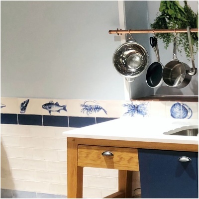 Kitchen corner with wooden blue cabinet, white countertop, hanging copper rod with pots, and marine-themed tile backsplash.
