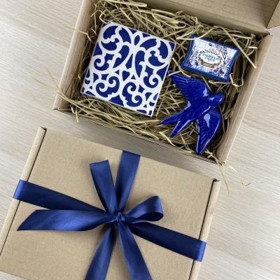 Gift box with blue ribbon containing blue and white ceramic tile, blue ceramic bird, and small labeled box.