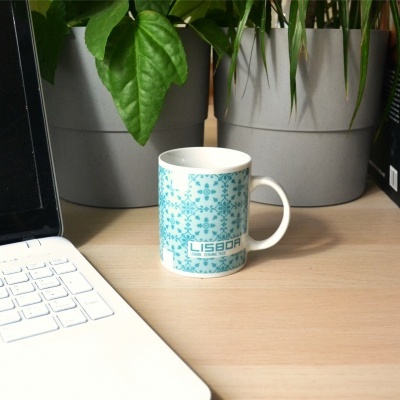 Caneca azul com padrão de azulejos ao lado de laptop, plantas em vasos e livros numa mesa de madeira