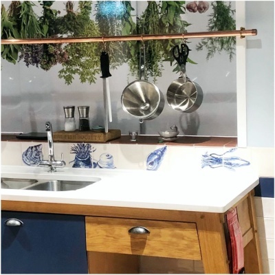 Kitchen countertop with white surface, wooden drawer, blue cabinet, stainless steel faucet, salt and pepper shakers, hanging herbs, pans and scissors on copper rod, blue sea creature tile illustrations