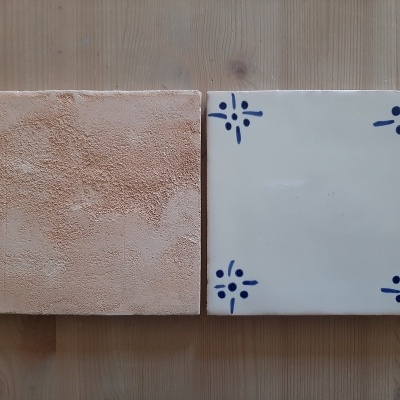 Dois azulejos quadrados, um bege e texturizado, outro branco com motivos azuis nos cantos, sobre madeira clara