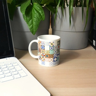 Caneca branca com padrão de azulejos coloridos numa mesa com computador, plantas e livros