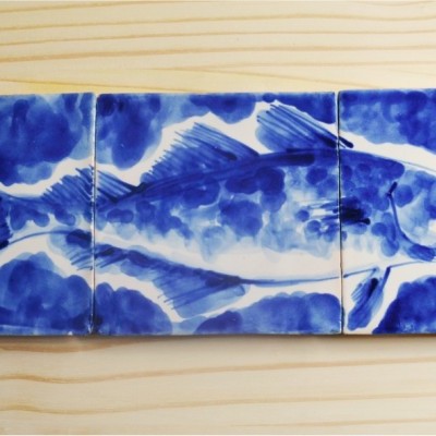 Três azulejos cerâmicos com pintura azul de um peixe em fundo azul sobre madeira clara