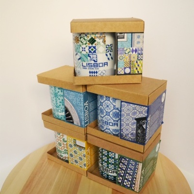 Caixas com canecas decoradas com padrões de azulejos e texto Lisboa num fundo branco sobre mesa de madeira.