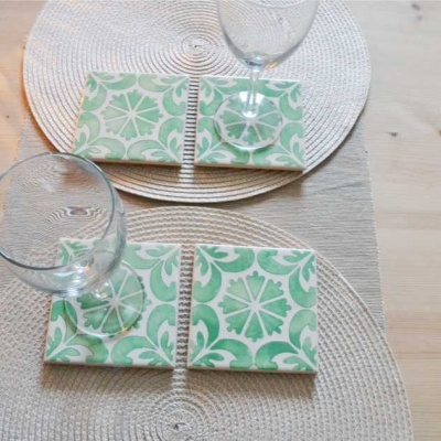Suportes de copos quadrados de cerâmica verde e branco com copos de vidro sobre toalhetes redondos bege numa mesa de madeira.