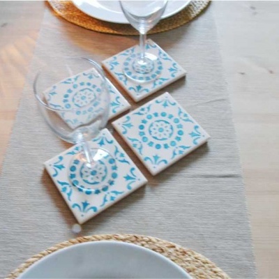 Quatro bases para copos quadradas com padrões florais azuis sobre uma mesa de madeira clara com toalha cinzenta.