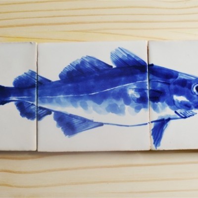 Três azulejos quadrados com desenho de peixe azul sobre mesa de madeira clara