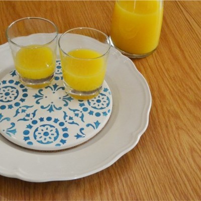 Dois copos com suco amarelo em tabuleiro azul e branco sobre prato branco numa superfície de madeira