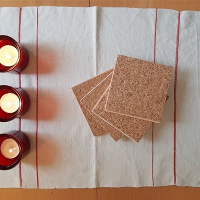 Três velas em copos vermelhos e bases quadradas de cortiça sobre pano branco com franjas.