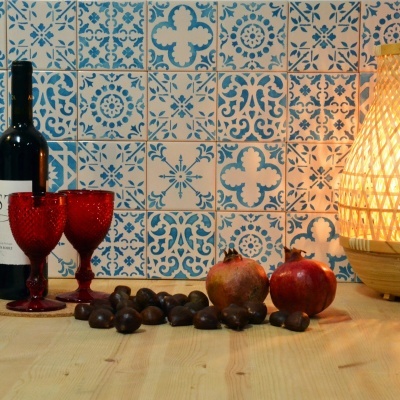 Mesa de madeira com garrafa de vinho, copos vermelhos, frutos e lâmpada de rattan na parede de azulejos