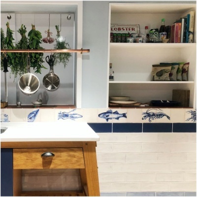Kitchen counter with hanging herbs and kitchen tools, blue and white seafood tile backsplash, and shelves with groceries and books