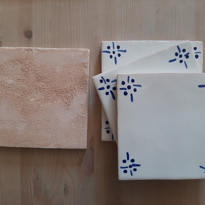 Quatro azulejos quadrados, um bege texturizado e três brancos com padrão azul nas extremidades, sobre madeira clara.