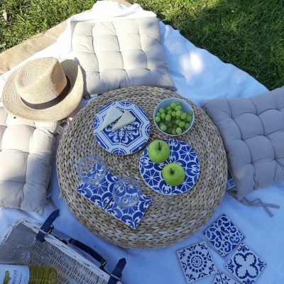 Mesa redonda de palha com pratos azuis e brancos, frutas, almofadas cinzentas, toalha branca, chapéu de palha e cesta de piquenique num campo de relva