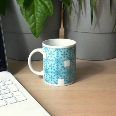 Caneca branca com padrão azul floral e geométrico sobre mesa junto a computador e plantas