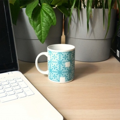 Caneca com padrão azul e branco sobre mesa de madeira ao lado de computador e plantas
