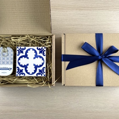 Cardboard gift box with chocolate sardines tin and decorative tile inside, next to a closed gift box with blue ribbon.