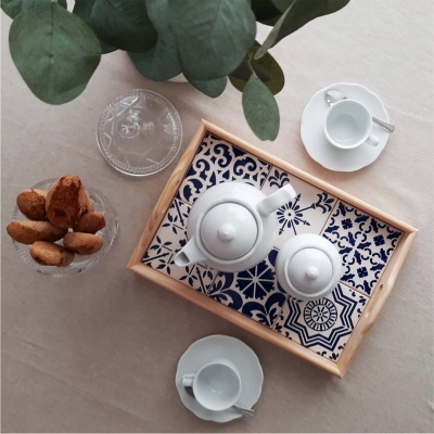 Tabuleiro decorado com azulejos azuis e brancos com bule e leiteira em porcelana branca, chávenas com pires e bolos numa taça 