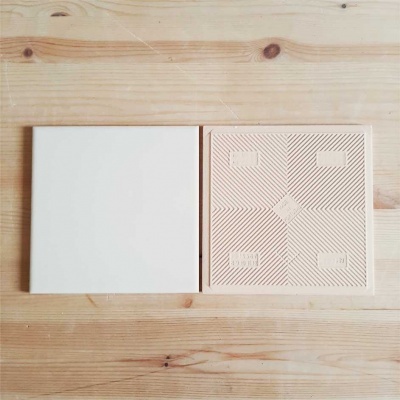 Dois quadrados, um branco e liso, outro bege com padrão geométrico e texto 'PARIS' em relevo, sobre superfície de madeira clara.