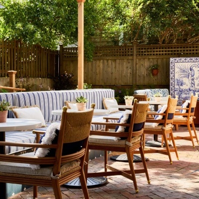 Área exterior com cadeiras em madeira e tecido, mesas brancas, sofá às riscas e painéis de azulejo azul e branco