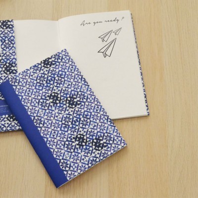 Dois cadernos com capa azul e branca com padrão geométrico, um aberto mostrando página com texto e desenhos de aviões de papel em mesa de madeira