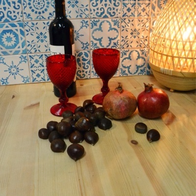 Mesa de madeira com vinho, taças vermelhas, castanhas, romãs e luminária ao fundo de azulejos azuis