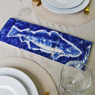 Mesa posta com jogos americanos, pratos brancos, talheres dourados e azulejos decorativos azuis com peixe.