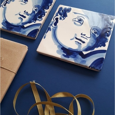 Dois azulejos azuis e brancos com faces humanas, fita dourada e objeto castanho sobre fundo azul.