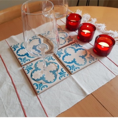 Quatro azulejos cerâmicos azuis e brancos sobre pano branco com franjas e linhas vermelhas, três velas vermelhas acesas e duas taças em mesa de madeira