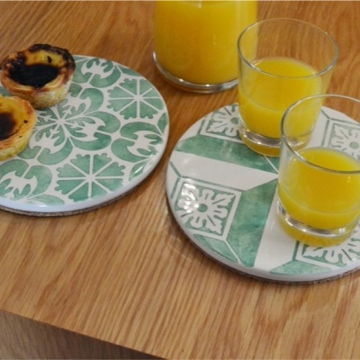 Dois pratos com padrão de azulejos verdes e brancos com pastéis de nata e copos de sumo de laranja sobre mesa de madeira.